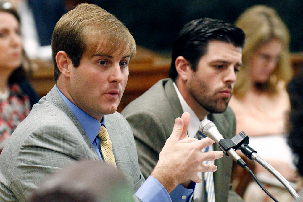 Two men in light grey suits speak into mics at a hearing