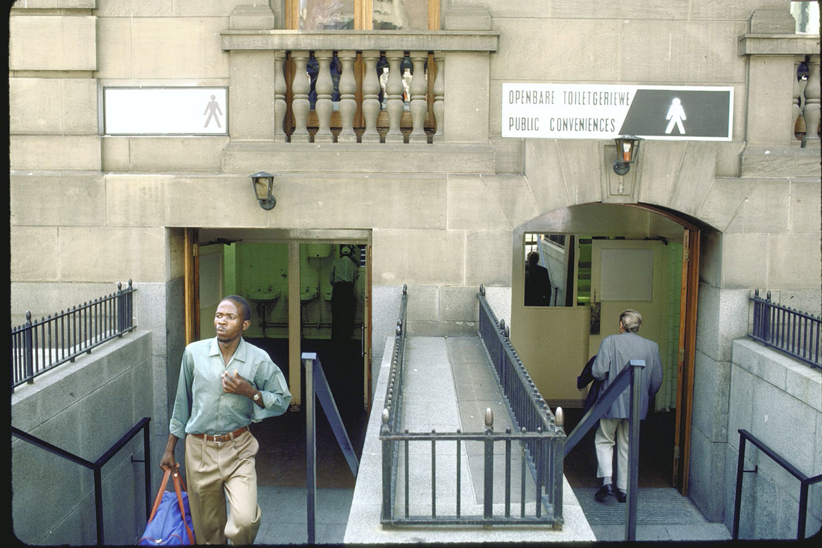 A Black man walks away from a limestone building, while a white man is seen entering on the other side. There are two signs above the entryways, one that shows a black man and the other shows a white man