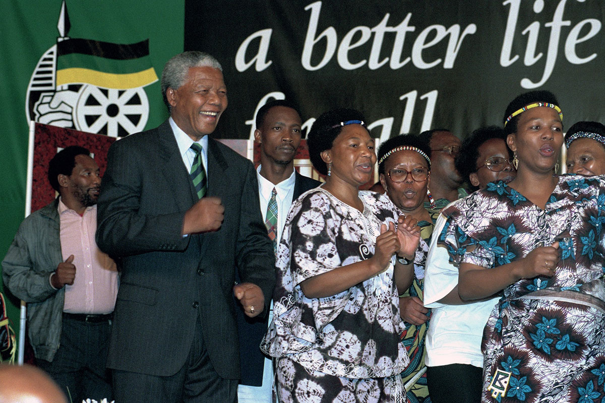 Nelson Mandela wears a dark suit and dances alongside women, in front of a sign that has the words 'a better life