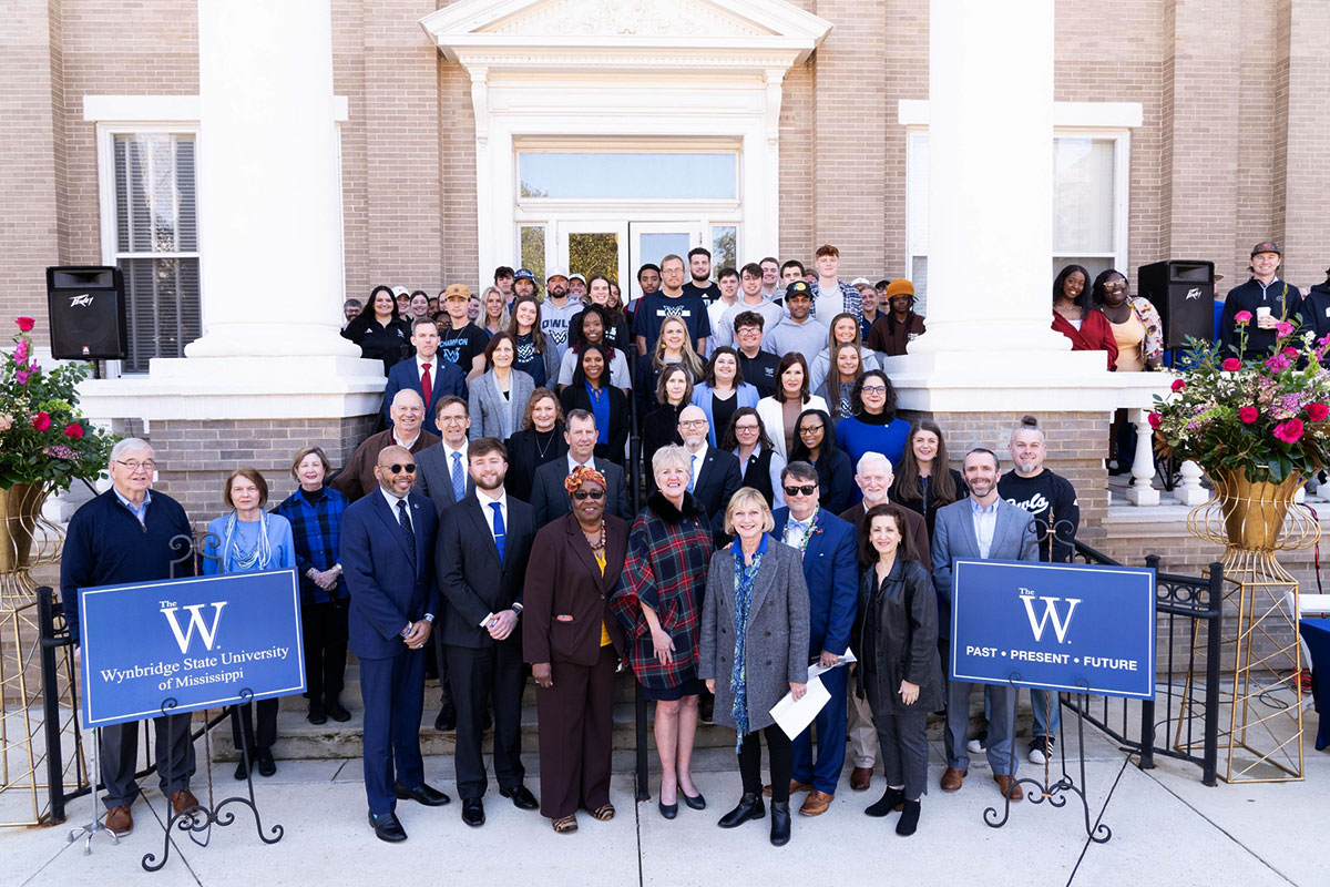 A large crowd of people gather in the steps of a brick building. Signs that read The W flank them.