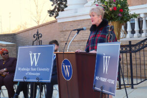 Mississippi University for Women President Nora Miller speaks at the podium. A sign beside her reads The W. Wynbridge State University of Mississippi
