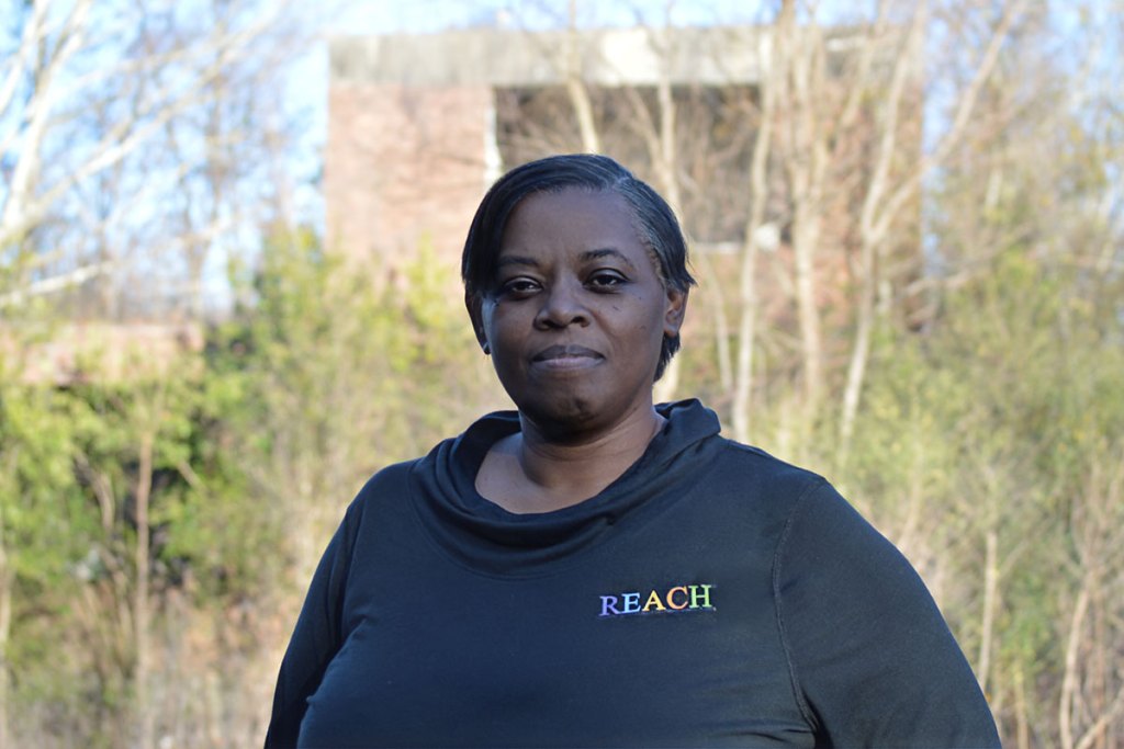 Putalamus White in a shirt that reads "REACH" stands outside with buildings faintly visible behind trees behind her