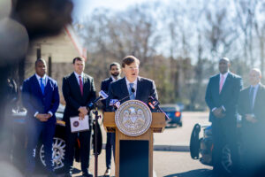 Gov. Tate Reeves standing at a podium outside of a gas station. Officers and men in suits stand behind him.