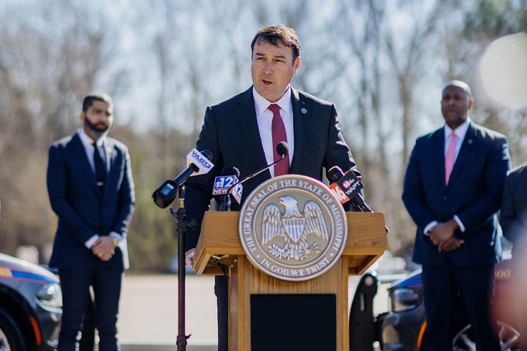 Commissioner Sean Tindall speaks at a podium labeled with a golden state seal of Mississippi