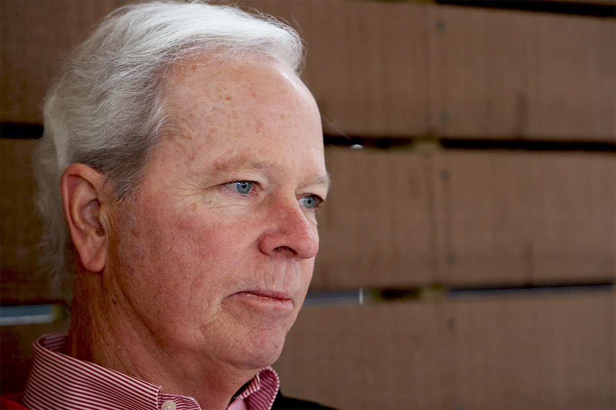 A closeup of an older man looking to the right of the frame. A brown wood slat wall is behind him.