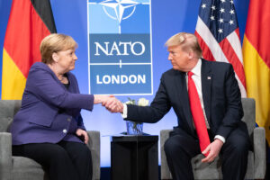 President Donald J. Trump and Chancellor of the Federal Republic of Germany Angela Merkel shake hands on stage. A sign behind them reads "NATO London"