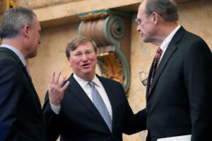 Three men in suits speak to each other in a building with beige marble walls
