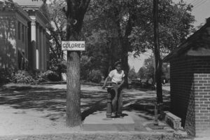 A young black boy is seen walking away from an outside water fountain. The tree beside it has a sign attached that reads "Colored"