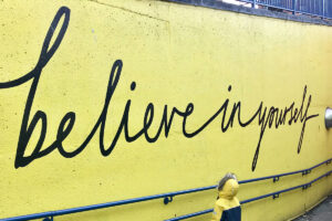 A small child looks up to a mural on a bright yellow wall that matches their coat - it reads "believe in yourself"