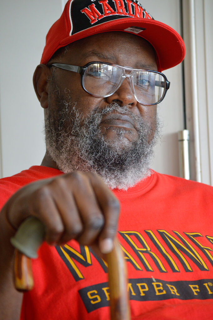 A seated man with walking cane in hand, wearing matching red Marines tshirt and cap