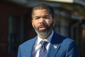 A photo of Mayor Chokwe A. Lumumba standing outside in a blue suit and tie. He's standing towards the sunlight and speaking