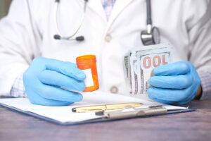 Close up of a doctor wearing blue gloves and lab coat, holding hundred dollar bills in one hand and a orange bottle of pills in the other