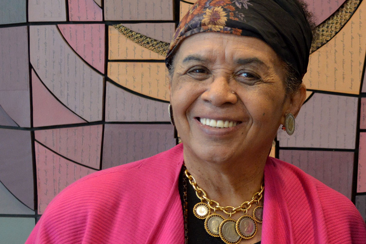 A woman in a pink top and black floral head scarf smiling in front of a colorful mosaic art piece on the wall