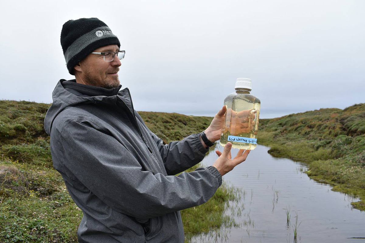A scientist in a rain jacket and cap holds up a water sample in a jar