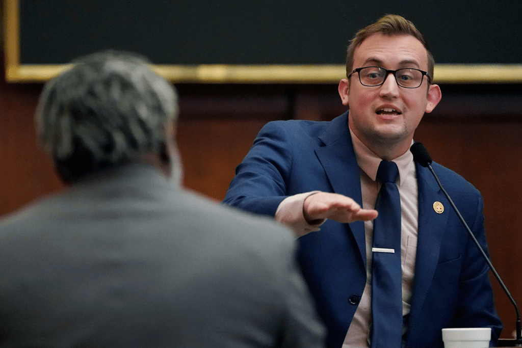 A man in a blue suit speaks at a mi and gestures to another man, who's back is to the camera