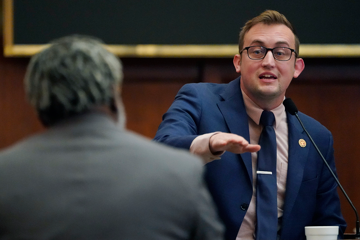 A man in a blue suit speaks at a mi and gestures to another man, who's back is to the camera