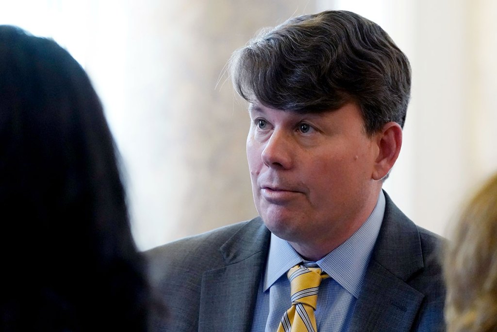 A closeup of a man in a grey suit with a blue striped yellow tie speaks in a crowd