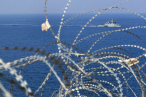 A view of a grey ship on the sea, as seen through a lot of barbed wire fencing