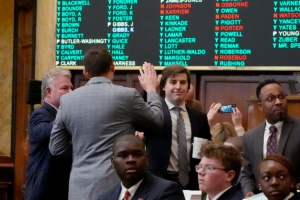 Two men in suits high five while other men look around. A voting result board with many names on it hangs behind them.
