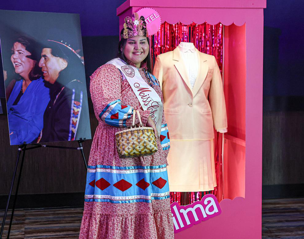 A woman wearing a Miss Cherokee sash over traditional dress smiles in front of a Barbie-style box that holds a creme colored two piece jacket and skirt set.