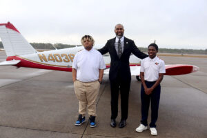 A man in a suit stands beside two younger men. A white and red airplane is behind them.