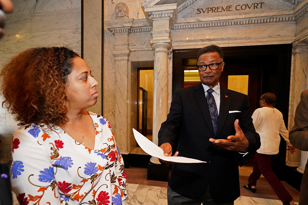 A man in a black suit proffers a piece of paper to a woman in a floral top. Behind them is a doorway labeled "SVPREME COVRT"