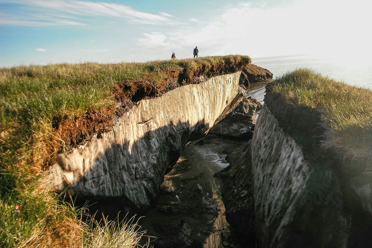 Rocky cliffs covered in grass have a split between them. The ocean can be seen beyond, as well as two people standing on the cliffs in the distance
