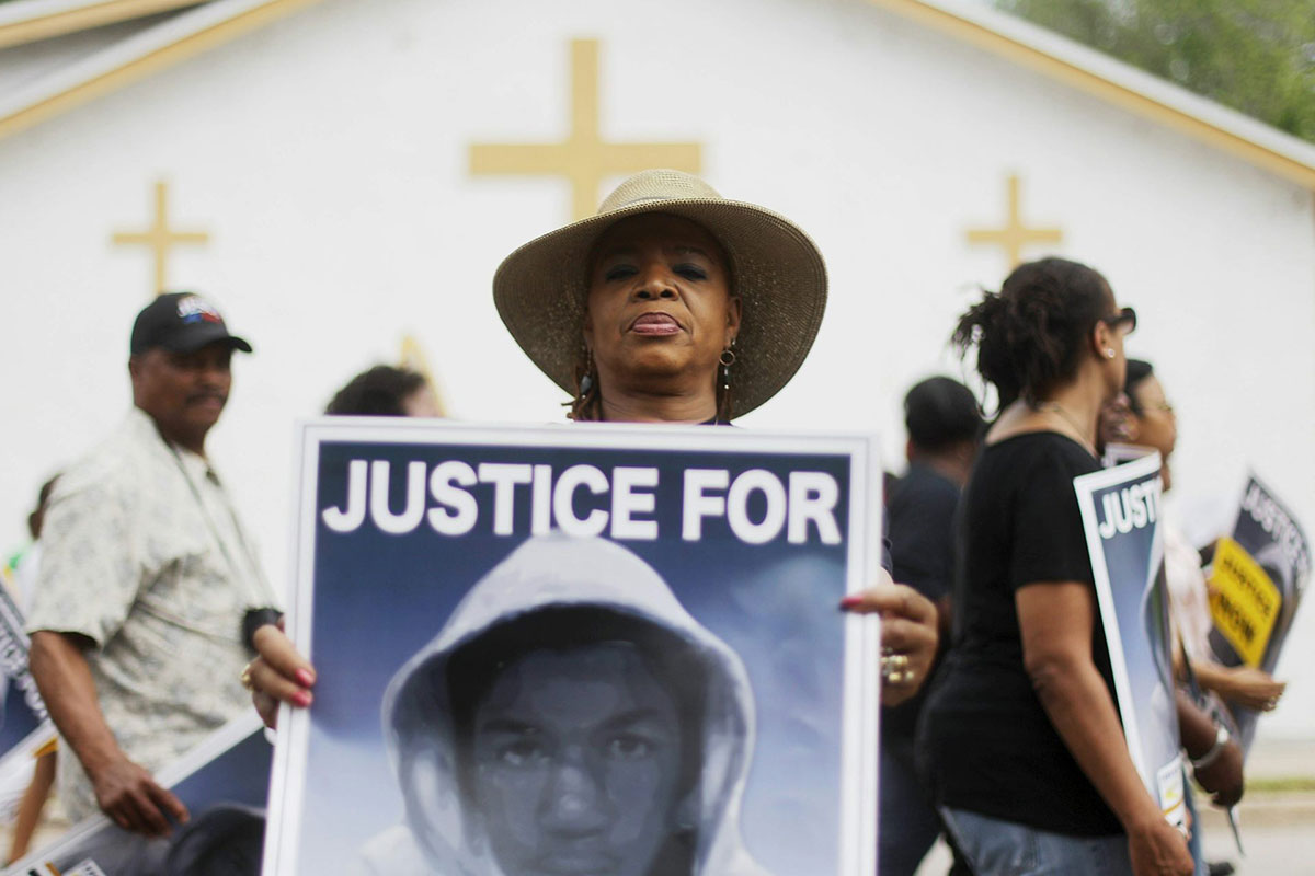 A Black woman stands in front of a church and holds a poster of a Black man wearing a hoodie.