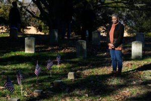 A woman stands in a shaded cemetery and looks onto a row of thirteen small square grave markers with US flags