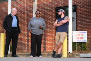 Three people stand in line outside of a red brick building waiting to vote. A sign to the right reads "Vote Here"
