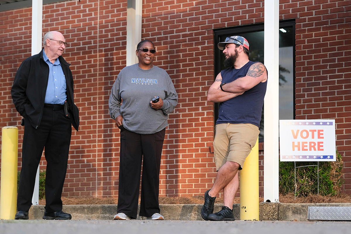 Three people stand in line outside of a red brick building waiting to vote. A sign to the right reads "Vote Here"