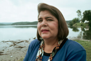 A woman with short brown hair and wearing blue clothing with a large beaded necklace stands outside on a shoreline