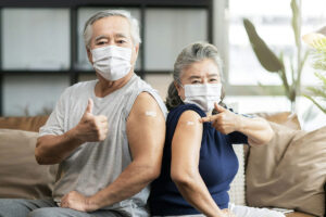 An older man and a woman, both wearing medical masks, point to their vaccine site bandages on their arms