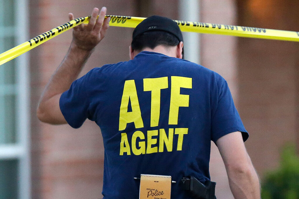 A man wearing a blue shirt with the words "ATF AGENT" in large yellow letters on the back is seen walking towards a crime scene, lifting the police line do not cross tape t o enter