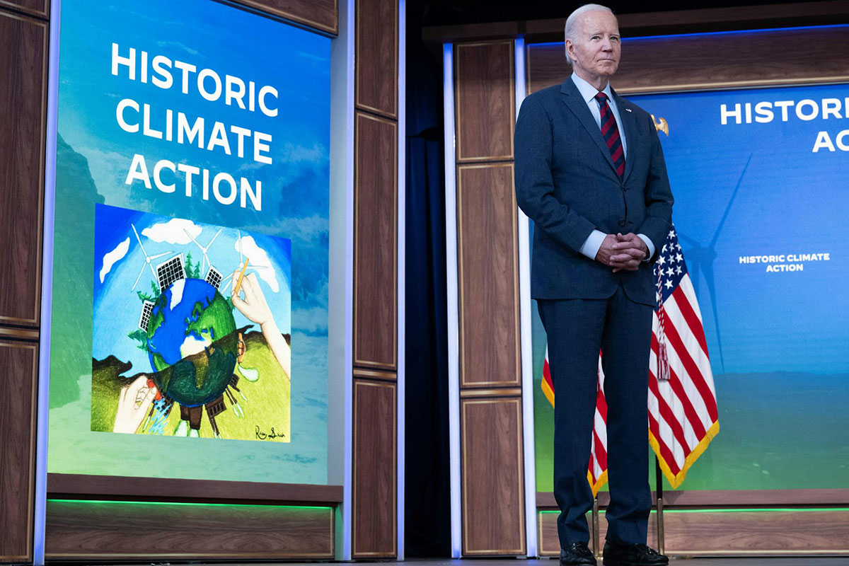 Joe Biden wears a blue suit and stands on a stage in front of a screen that says 'historic climate action (climate change)