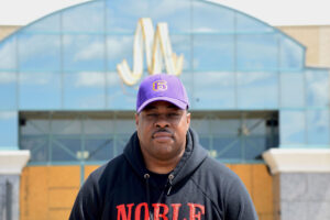 Man in a black hoodie and purple baseball cap with the number 6 on it stands in front of boarded doors to the Metrocenter Mall