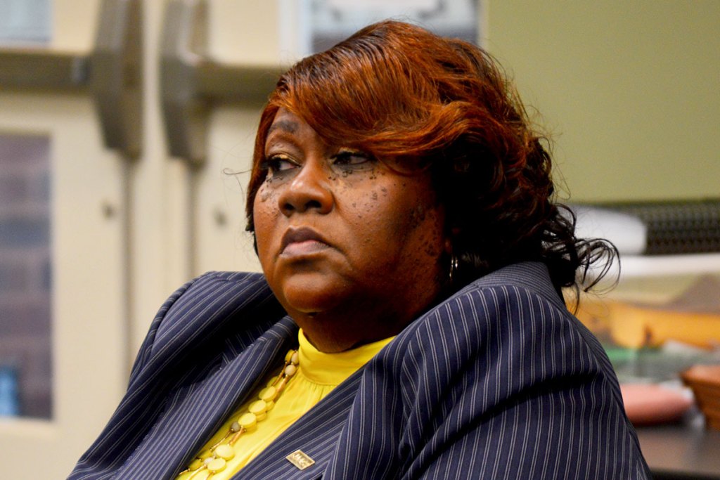 A closeup of a woman in a yellow top and pin striped jacket sits pensively