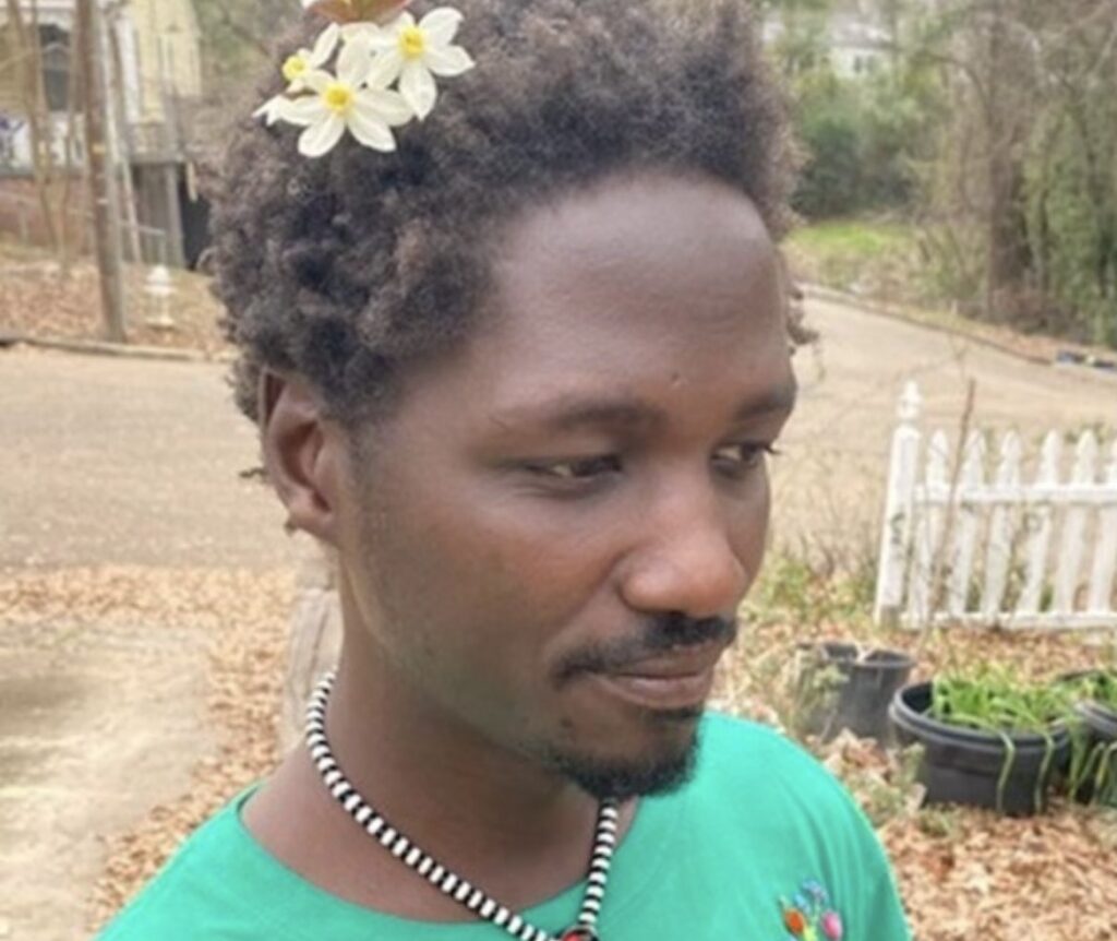 Closeup of a man in a green tshirt and a black and white necklace, with white and yellow flowers sitting in his short brown hair