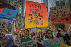 Young people carry many signs as they march down a road lined with tall buildings. Examples of signs "We're to Blame. The Earth is Aflame." "Biden: Our Future is on Fire" "Hot Girl Summer Not Sweaty Girl Summer" (climate change)