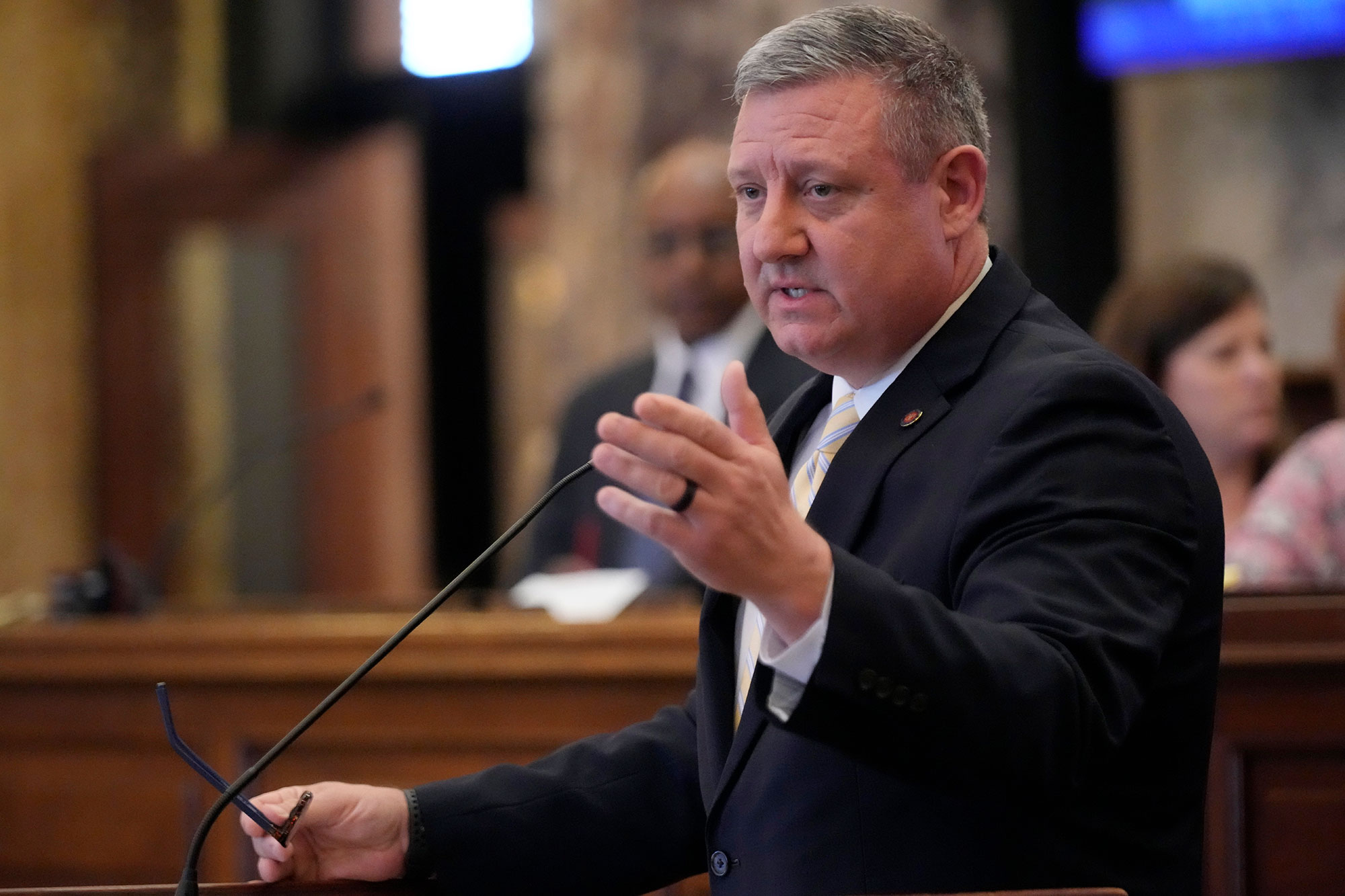 A senator in a black suit speaks at the podium, glasses in one hand