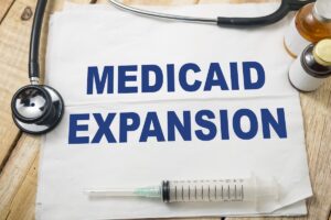 A piece of paper with the words MEDICAID EXPANSION printed in blue sits on a wooden surface, surrounded by a syringe, medicine bottles, and a stethoscope