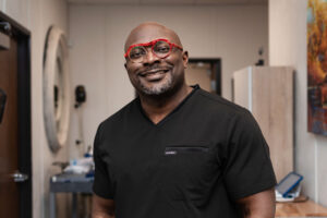 A man wearing black medical scrubs and round red glasses smiles inside a clinic hallway
