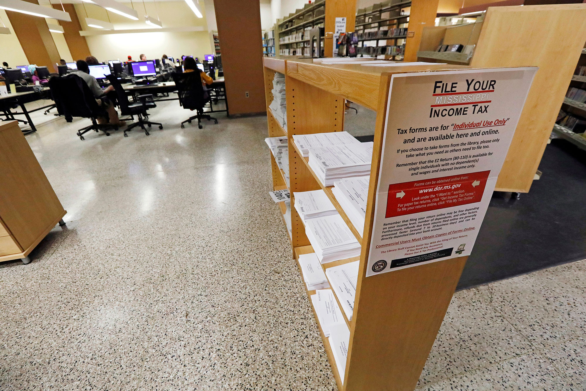 a view inside of a library. Shelves closest have Mississippi tax paperwork on them. Further back are tables with computers, many people sitting at them, with rows of bookshelves to the right