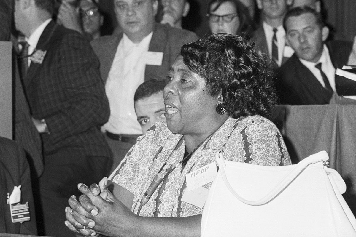 A woman sits at a table, speaking to a crowd of men around her in a meeting