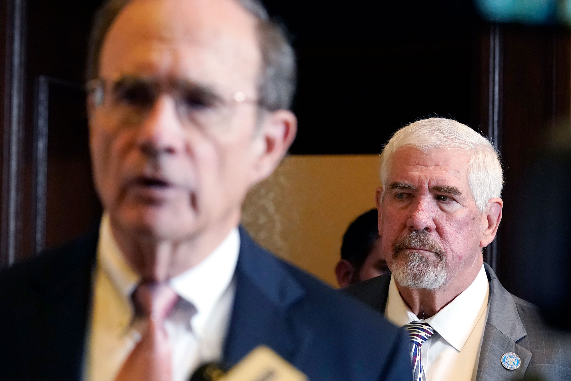 A man in a grey suit, fully in focus, stands behind and listens as a man in a navy suit, out of focus in the foreground, speaks