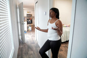 A woman in black and white clothes stands in front of a bedroom in a renovated three-bedroom trailer