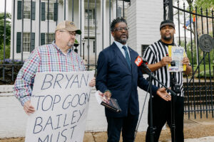 A man in a dark suit speaks at a mic in front of the governor's mansion. Men stand on both sides of him, holding signs.