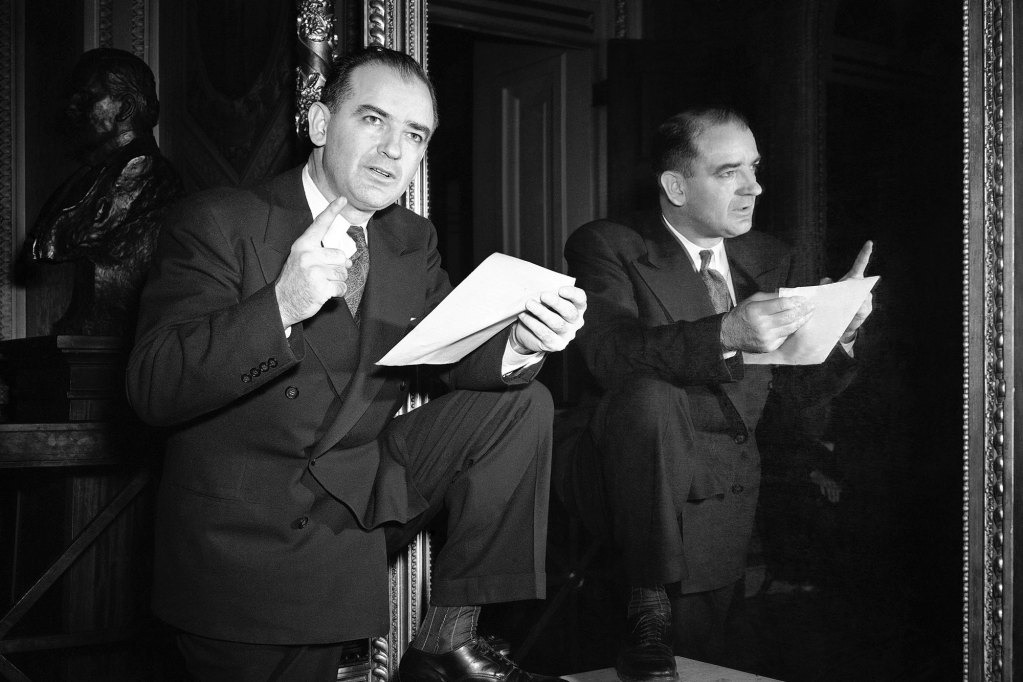 A black and white photo of a man in an 1950's style suit posed in front of a mirror. He has one foot propped on a low table, and is holding a piece of paper in one hand and pointing up with his other hand