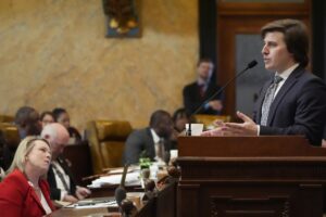 Kent McCarty standing at a microphone in front of the mississippi House floor while other representatives watch and listen while he explains school funding