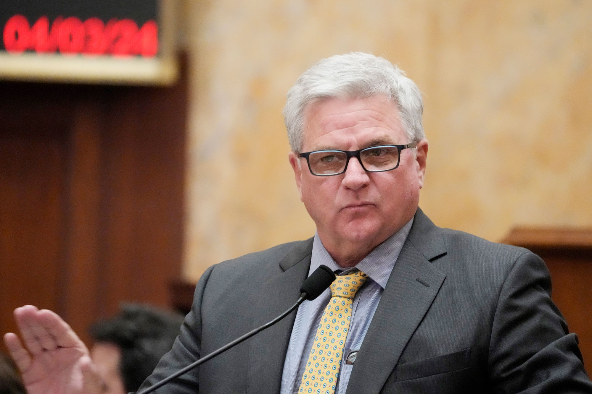 A man in a grey suit and yellow polkadotted tie holds his hand up in a "stop" gesture while speaking at a podium